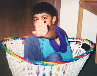 A young boy sitting in a rainbow-edged basket, smiling with his hand raised.