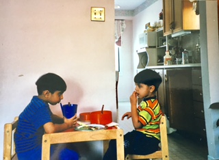 Two boys sitting at a small table in a kitchen, each doing their own activity.